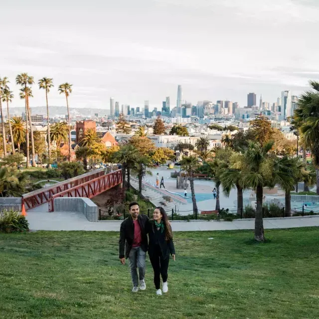 Um casal caminha em direção à câmera com Dolores Park e o horizonte de 威尼斯人官网平台app atrás deles.